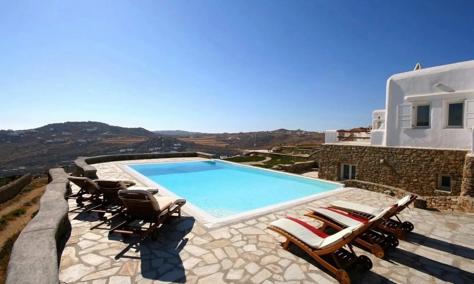 A swimming pool and sun loungers sit on a stone terrace overlooking hills.