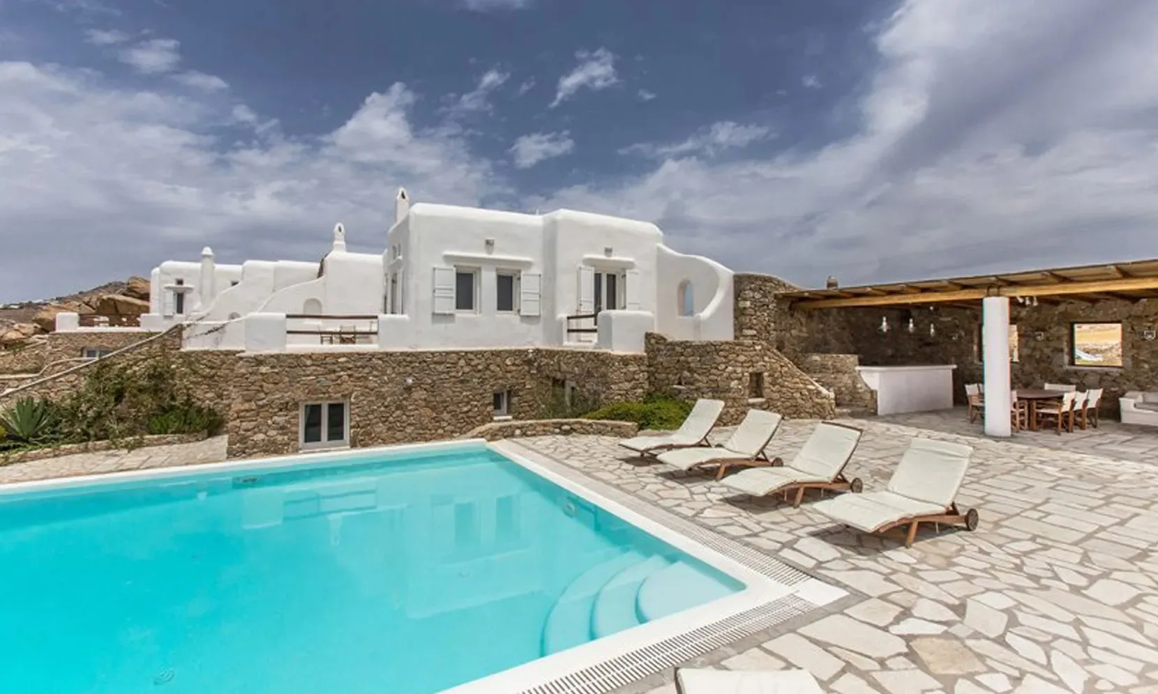 An outdoor swimming pool sits beside a stone terrace and white buildings.