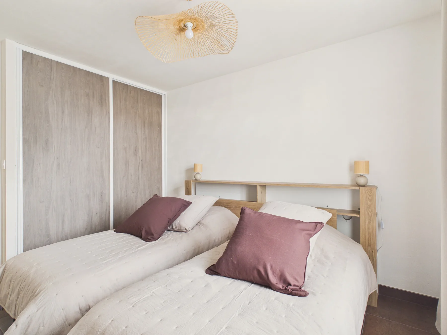 A bright bedroom with two beds, neutral bedding, and mauve accent pillows.