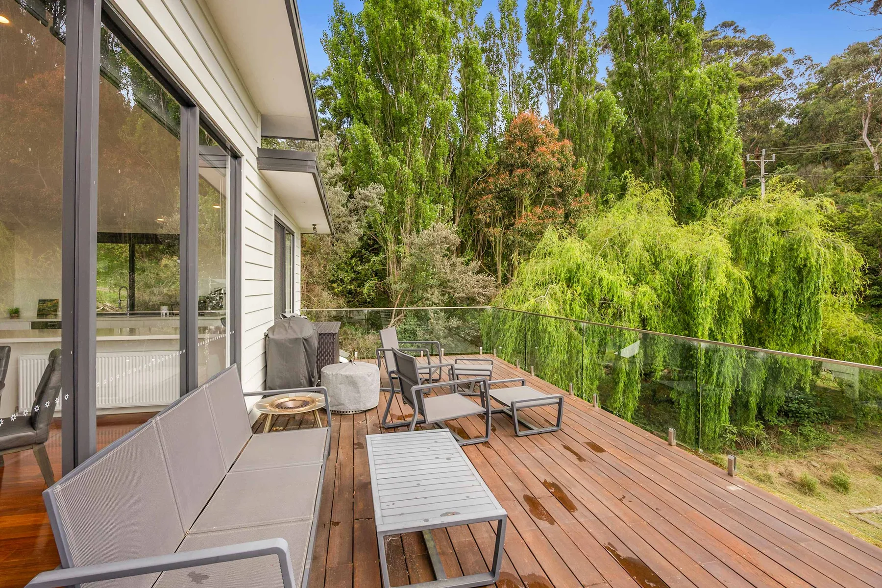 Una terraza exterior junto a una casa con paredes de vidrio tiene asientos y vistas a la vegetación.