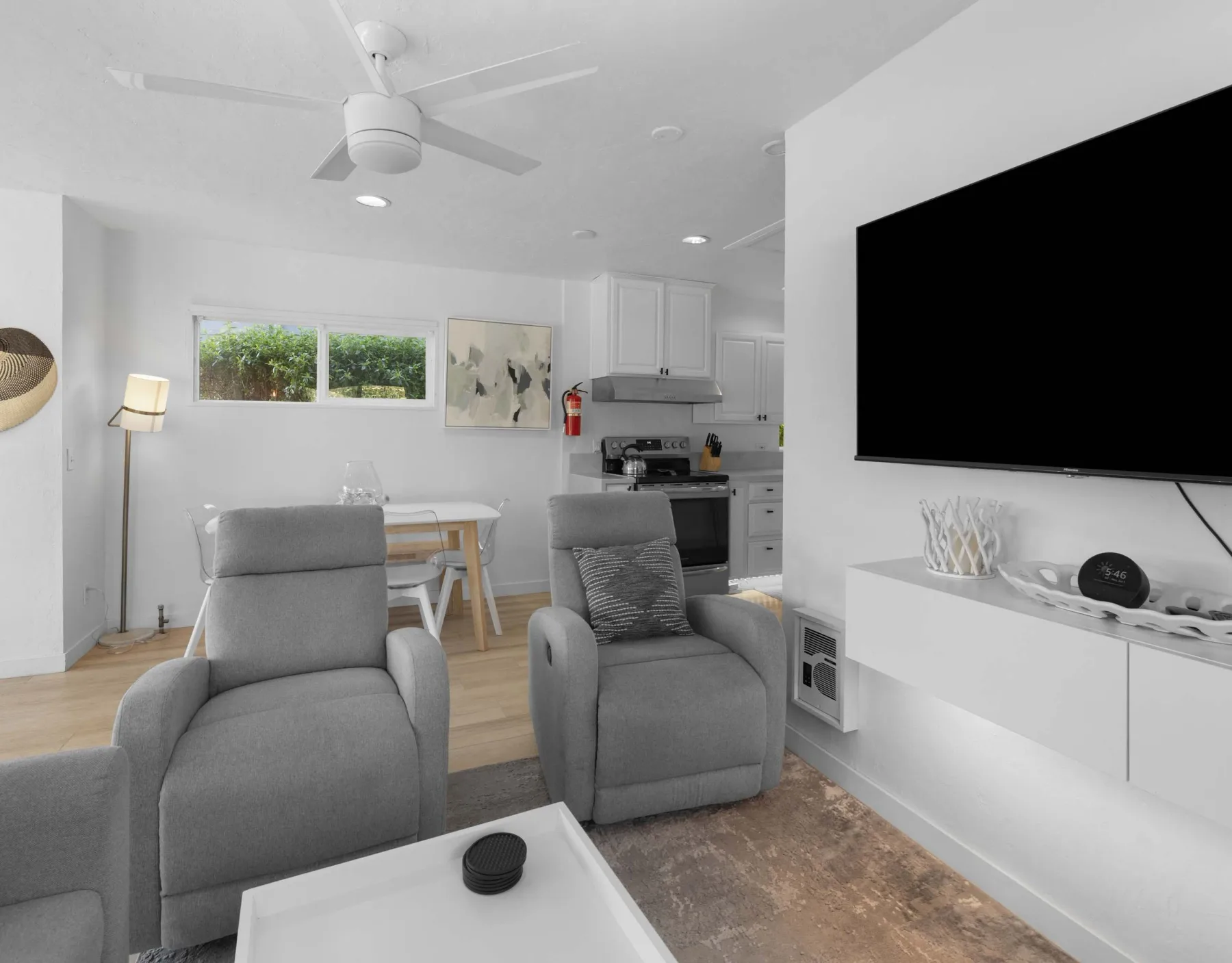 A bright living area with gray seating, a wall-mounted television, and an open kitchen.