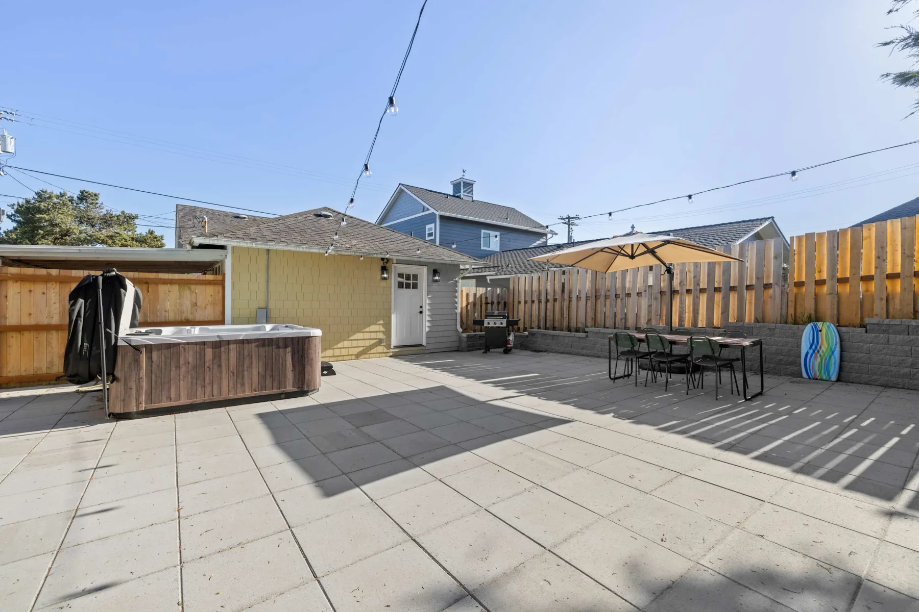 A paved outdoor patio has a hot tub, dining table, and wooden fence.