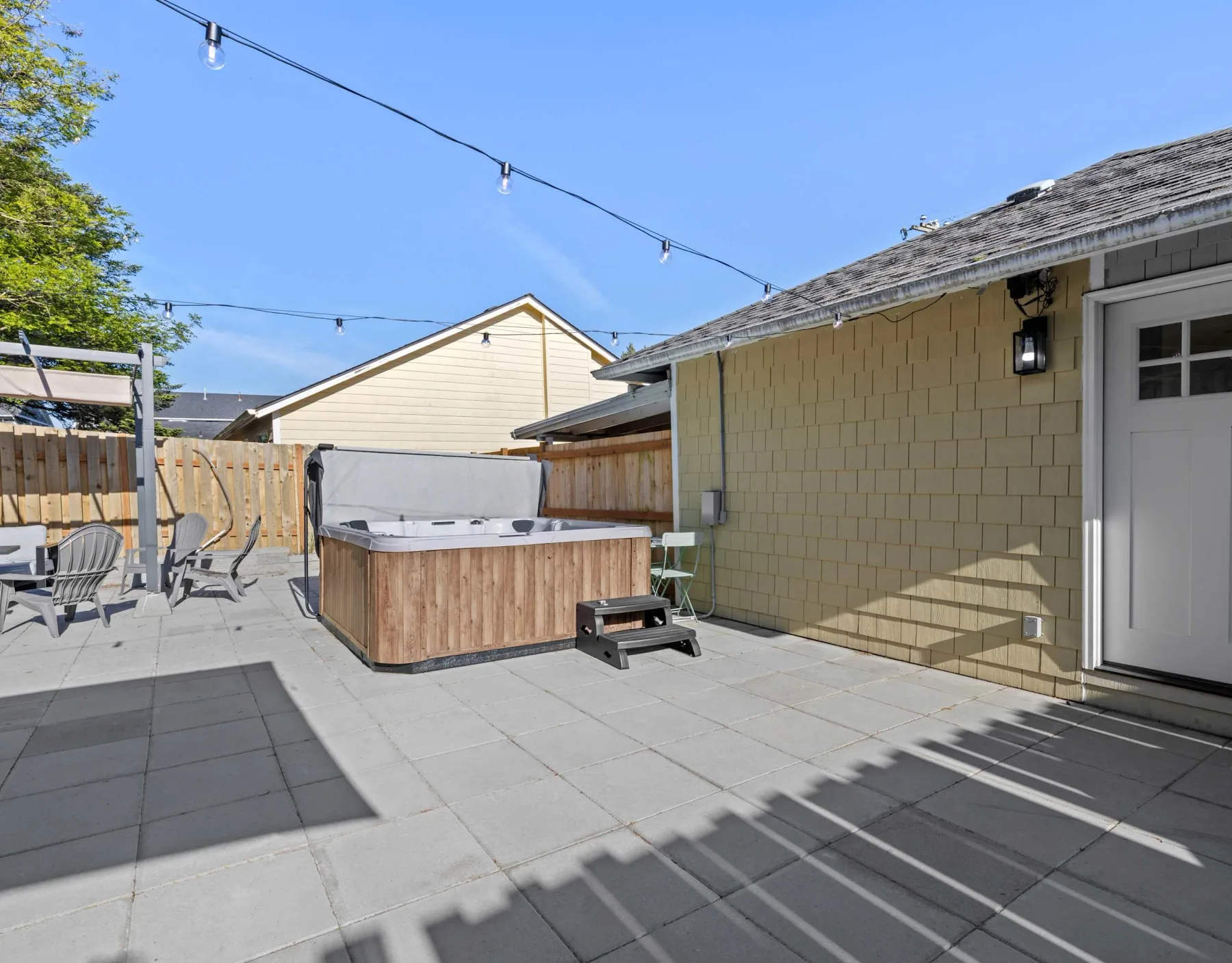 A paved outdoor patio with a hot tub, seating, and string lights.