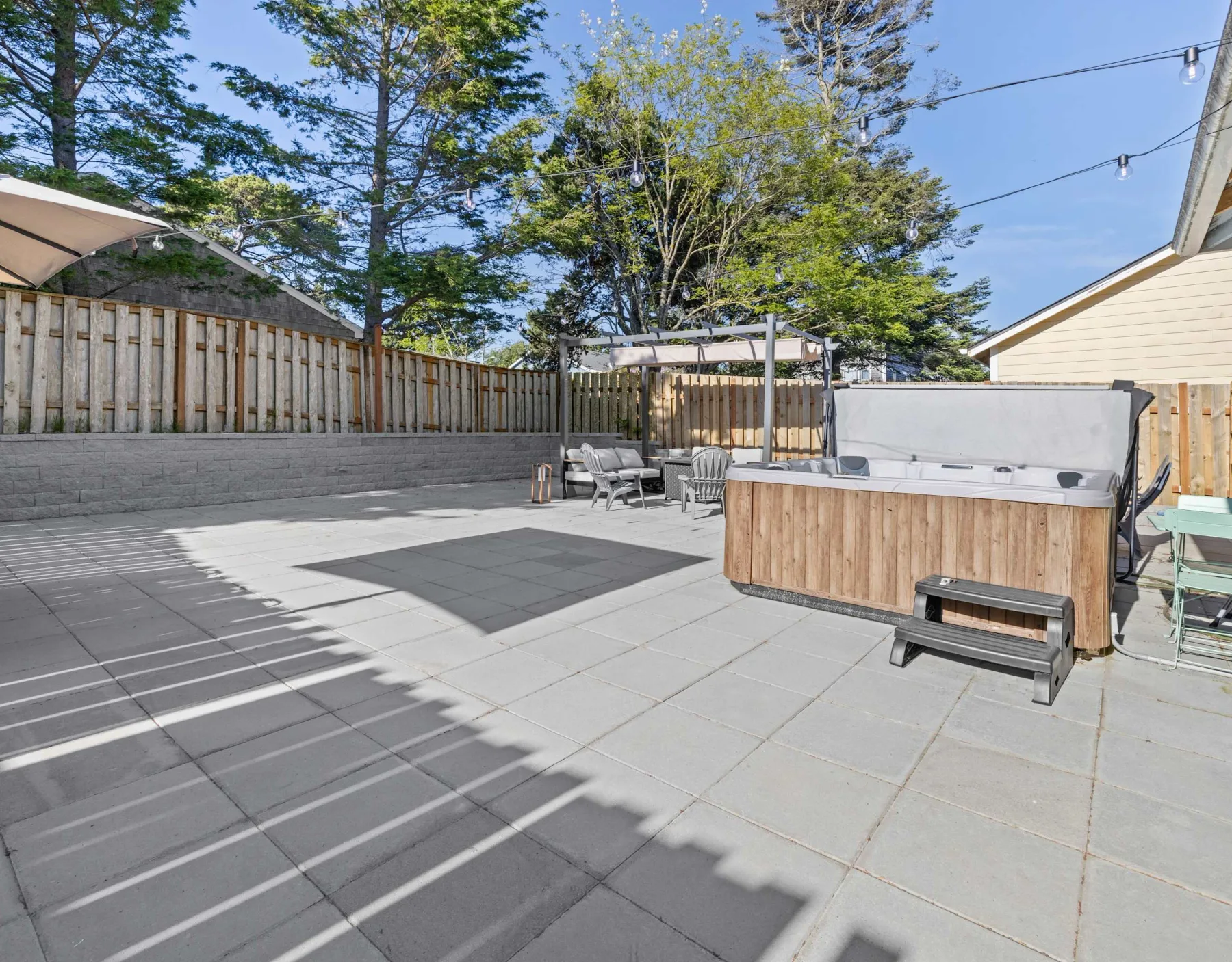 A paved outdoor patio with a hot tub and seating beneath the trees.