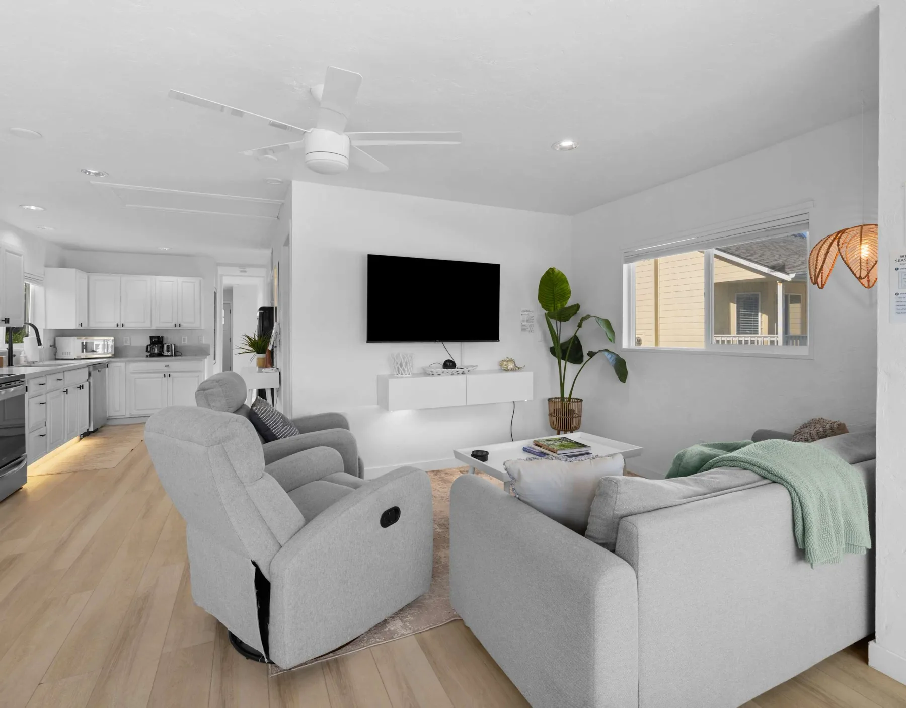 A bright living room opens into a white kitchen, with gray seating and a wall-mounted television.