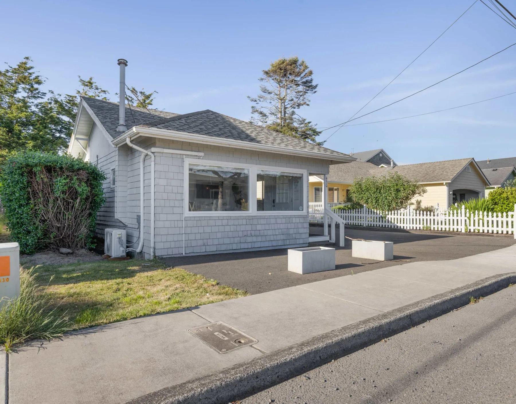 A small gray house sits beside a sidewalk, with a white picket fence nearby.
