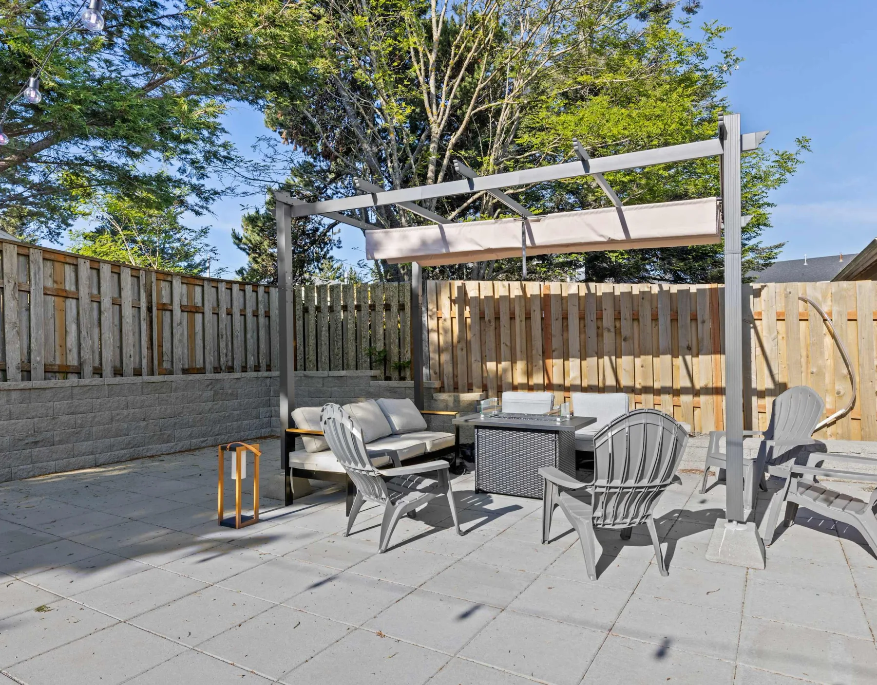 A paved outdoor patio with seating beneath a pergola, enclosed by a wooden fence.
