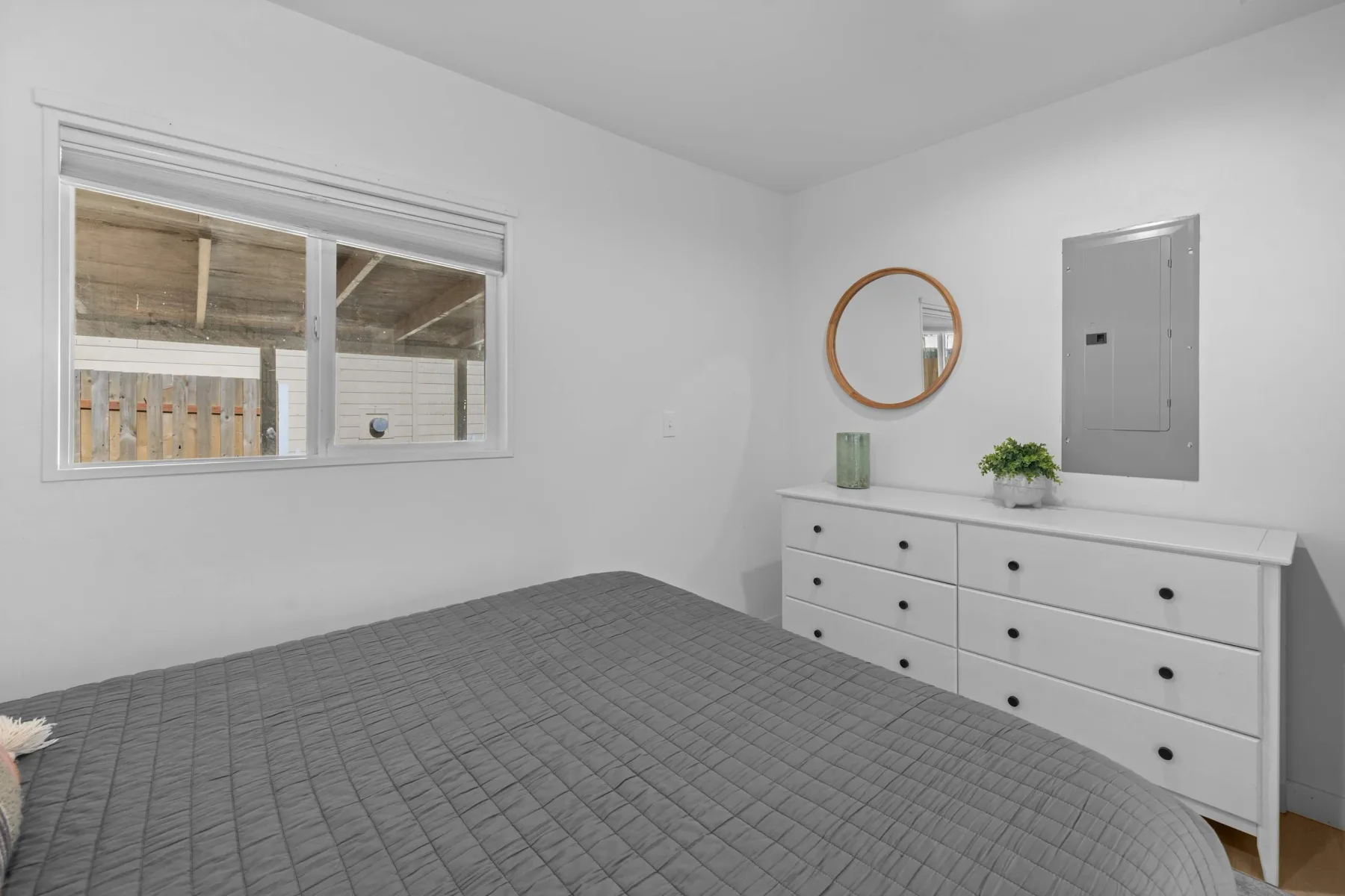 A bedroom with a gray bedspread, white dresser, and round wall mirror.
