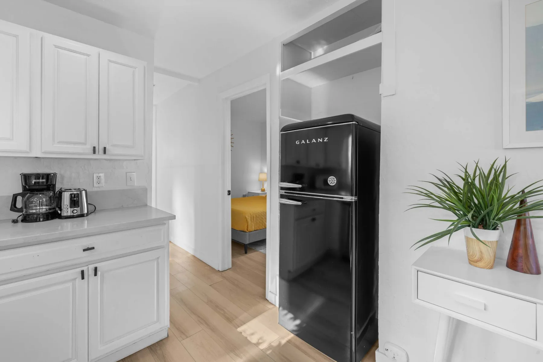 A bright kitchenette with white cabinets, a black refrigerator, and a small dining area beyond.