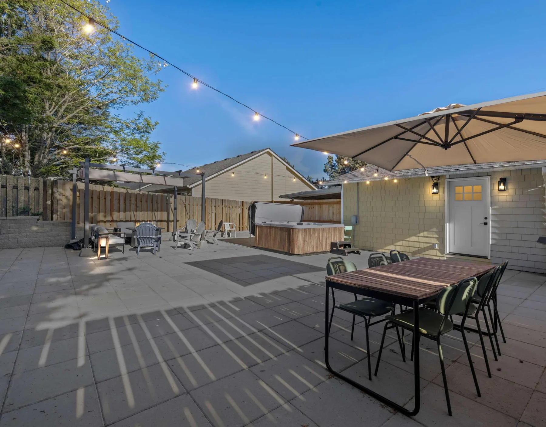 An outdoor patio with a dining table, seating, and a hot tub beneath string lights.