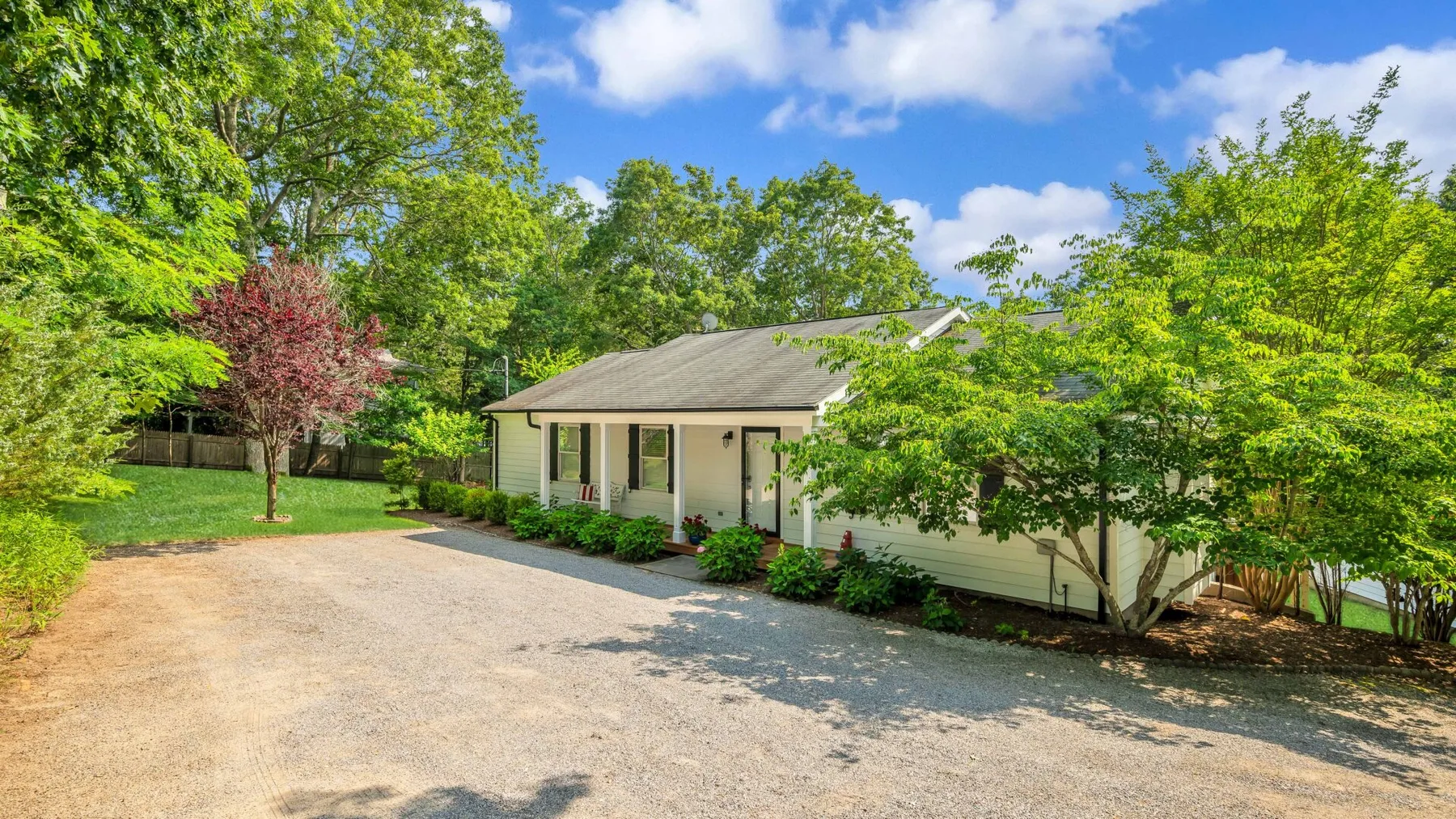 A single-story house stands beside a gravel driveway, surrounded by trees.