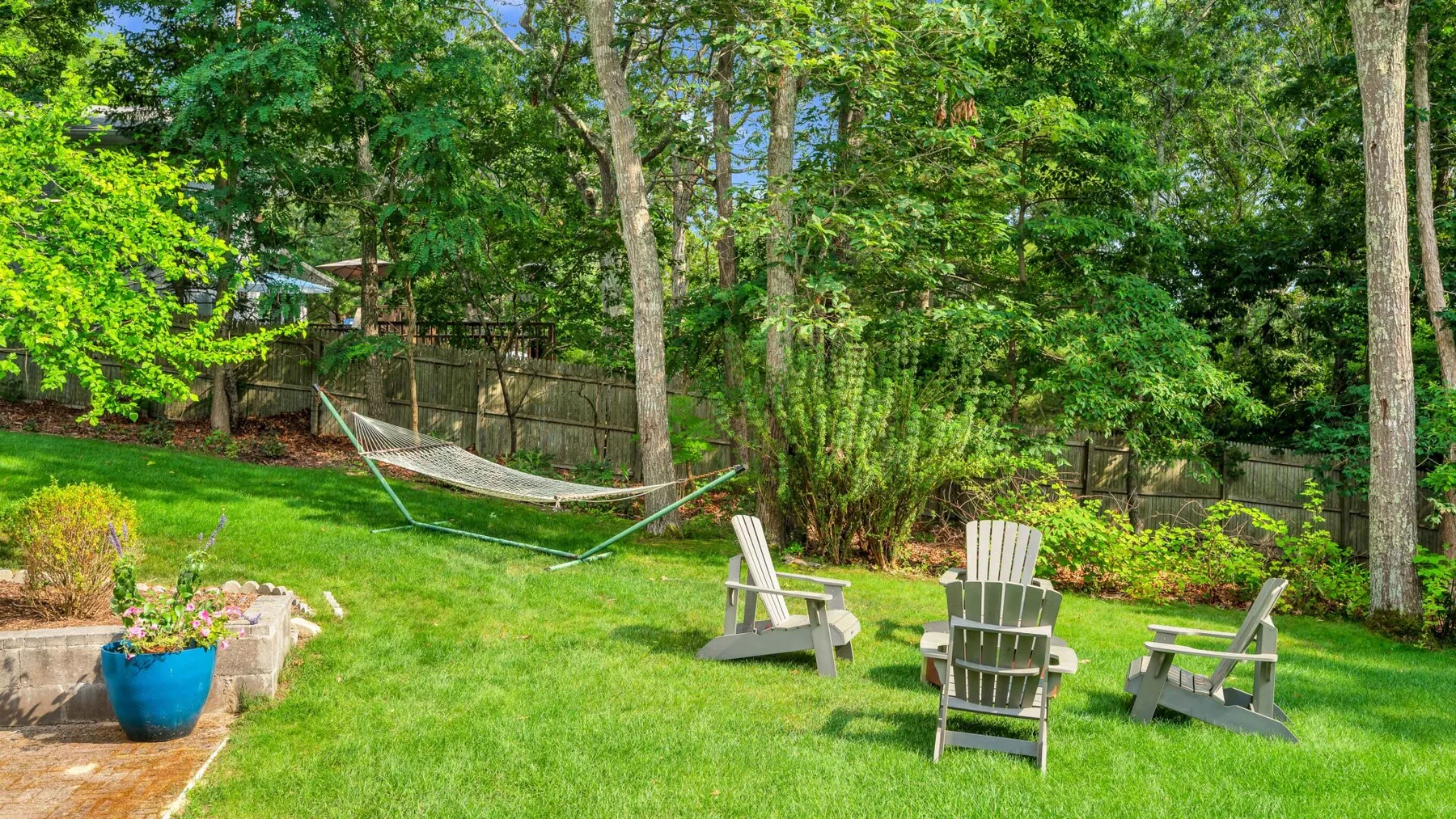 A leafy garden with a hammock and Adirondack-style seating on the lawn.
