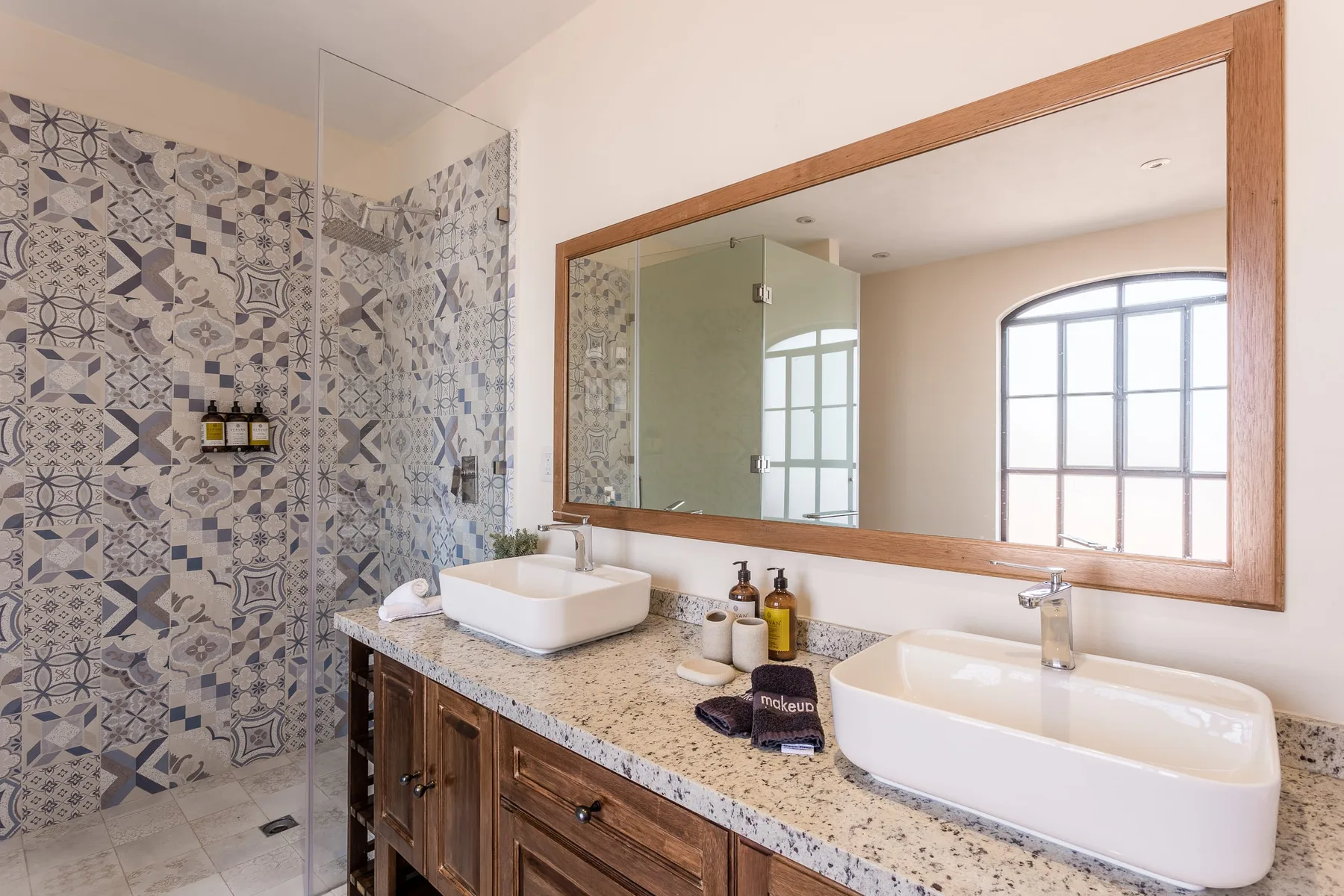 Un tocador de baño con dos lavabos blancos está junto a una ducha de azulejos estampados.