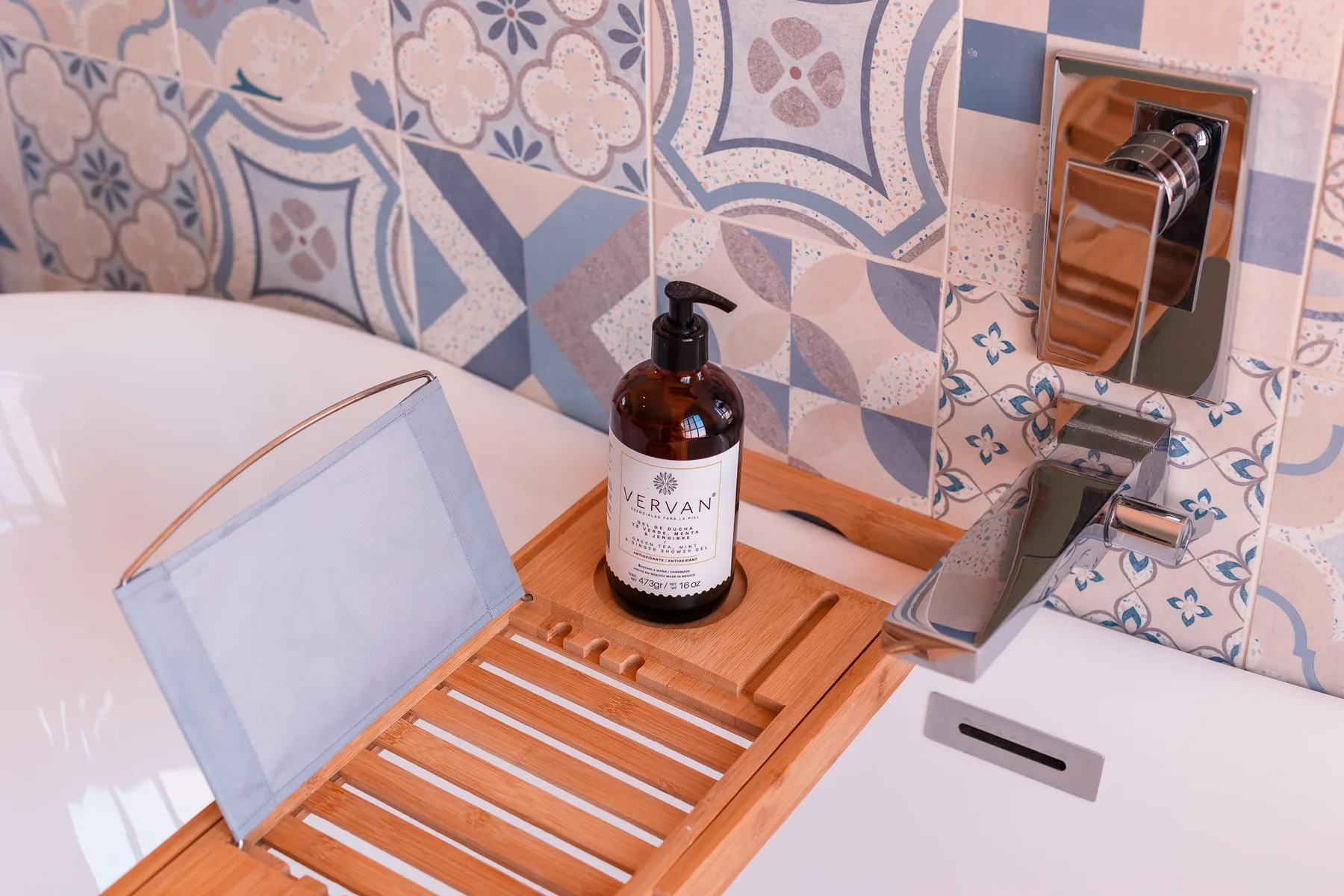 Una bañera con una bandeja de madera y un frasco de jabón está junto a azulejos estampados.