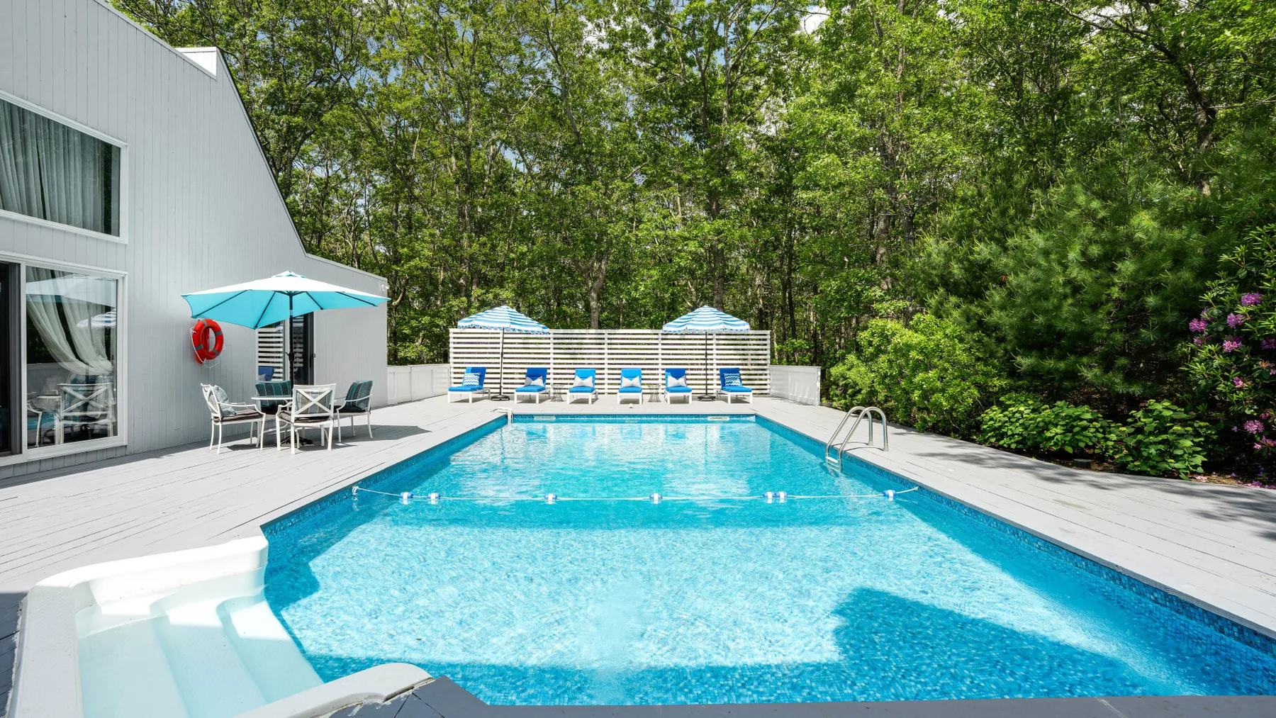 An outdoor swimming pool beside a white house, with lounge seating and trees.