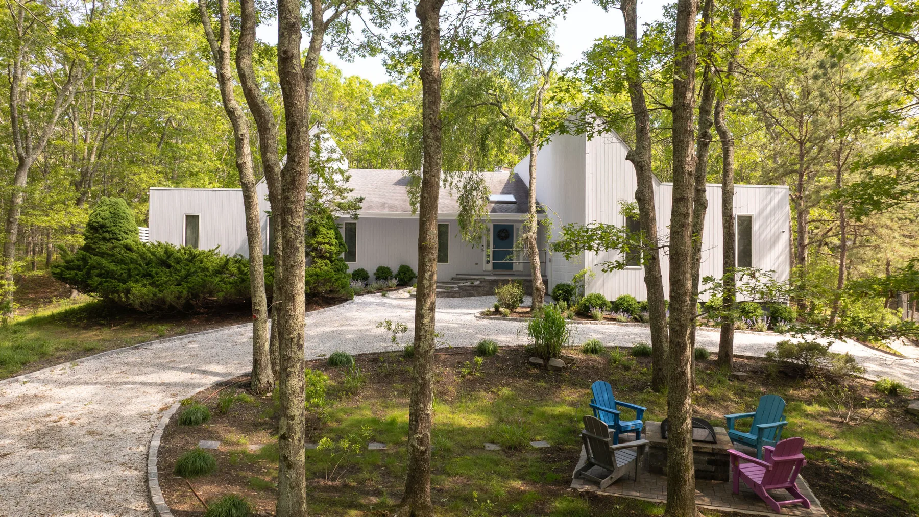 A modern house sits among trees, with a curved driveway and outdoor seating.