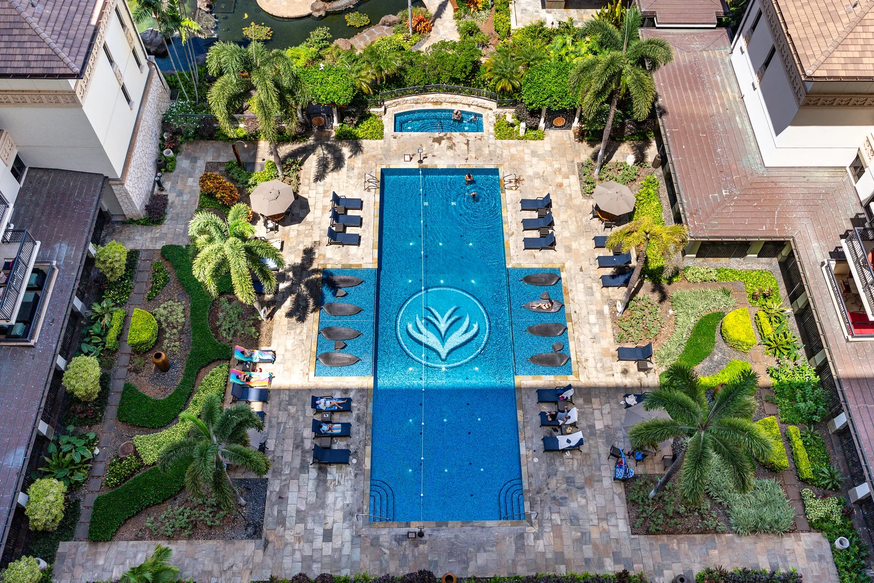 Vista aérea de una piscina rodeada de zonas pavimentadas con asientos y vegetación tropical.