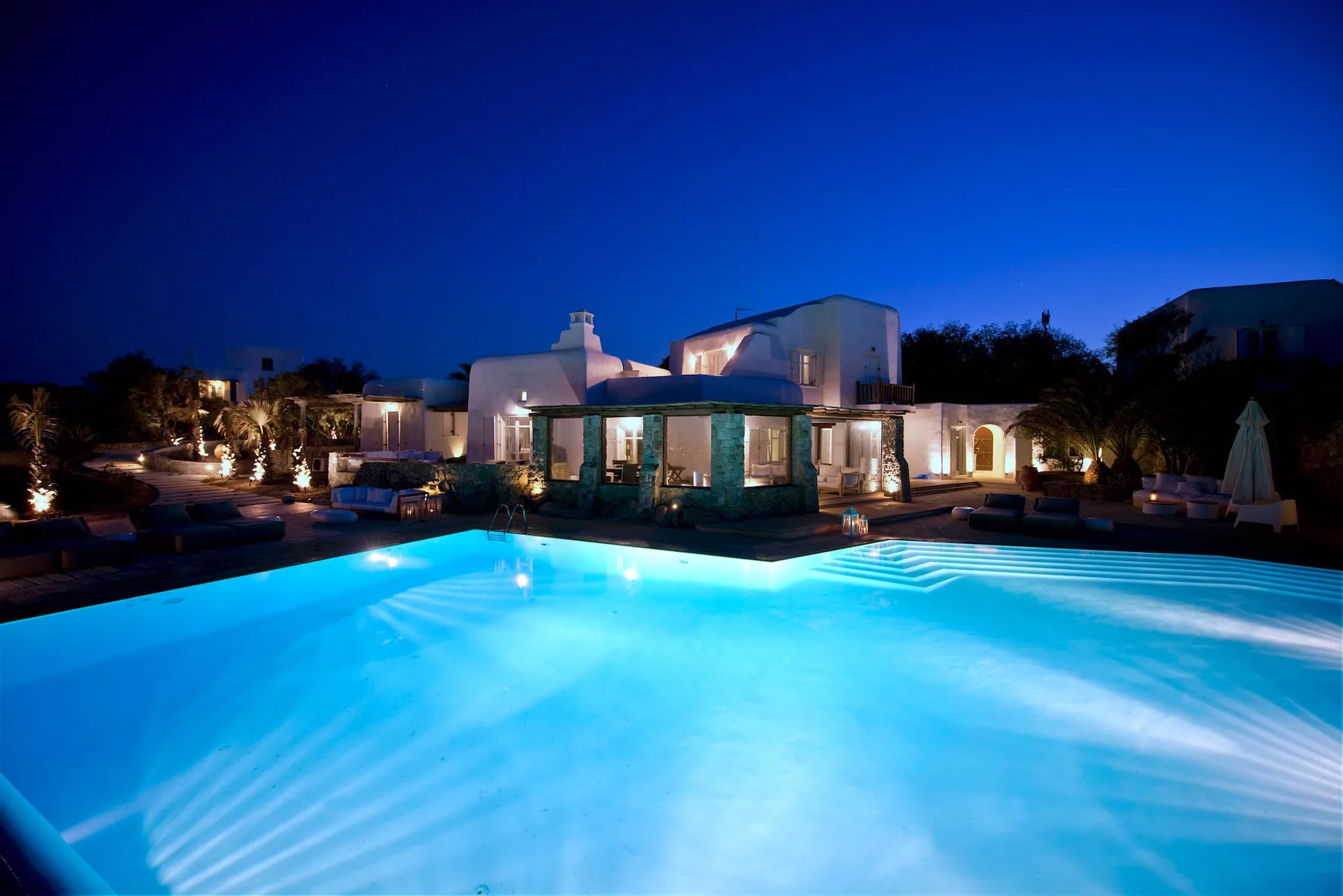 An illuminated swimming pool sits in front of a low building at dusk.