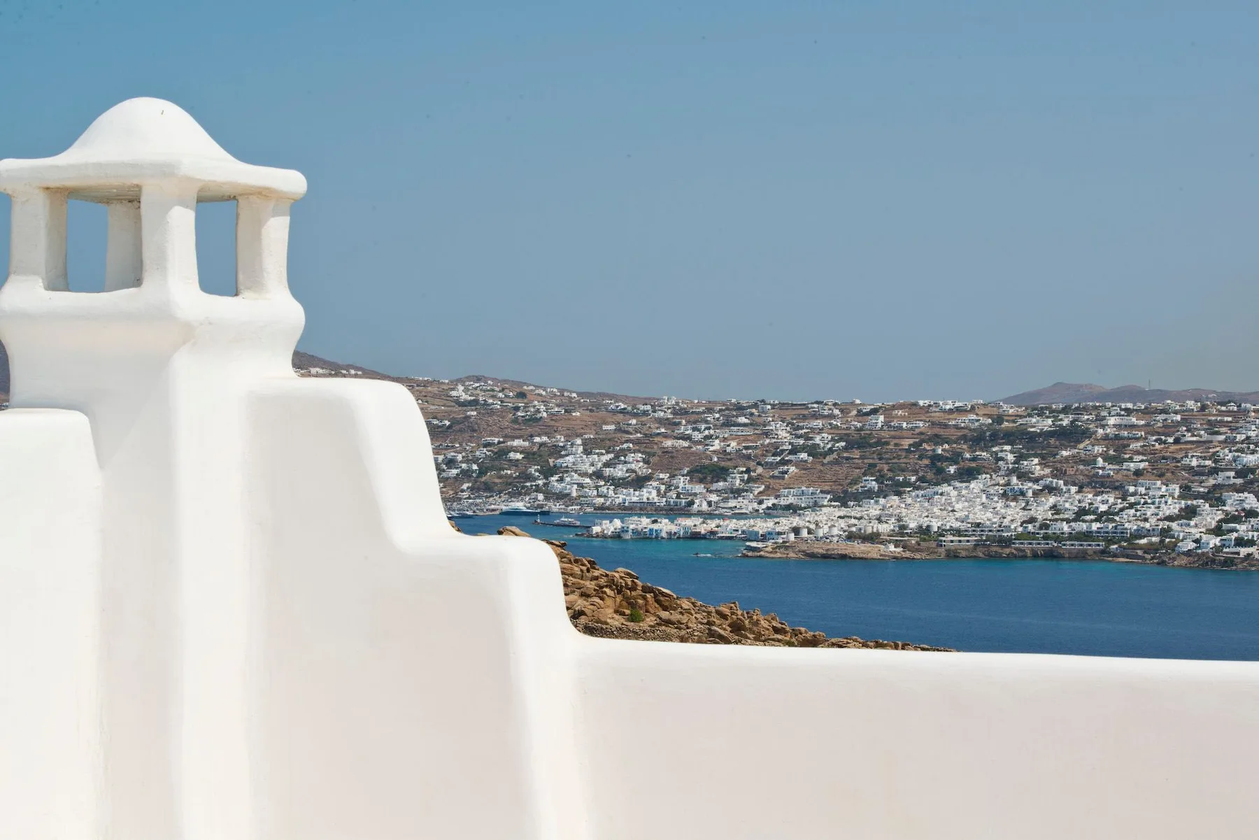 A white building facade overlooks a blue bay and a densely built hillside.