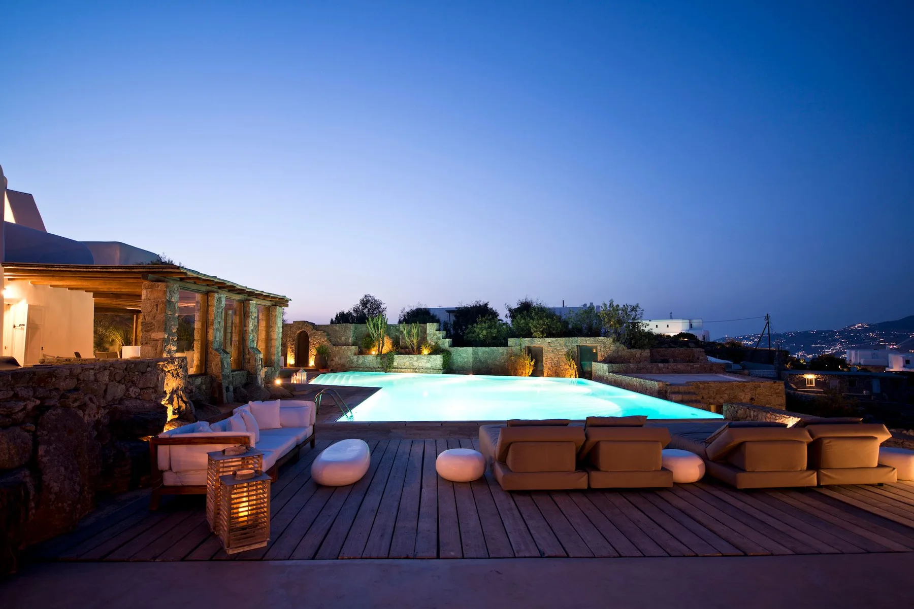 An outdoor pool glows beside lounge seating on a wooden deck at dusk.