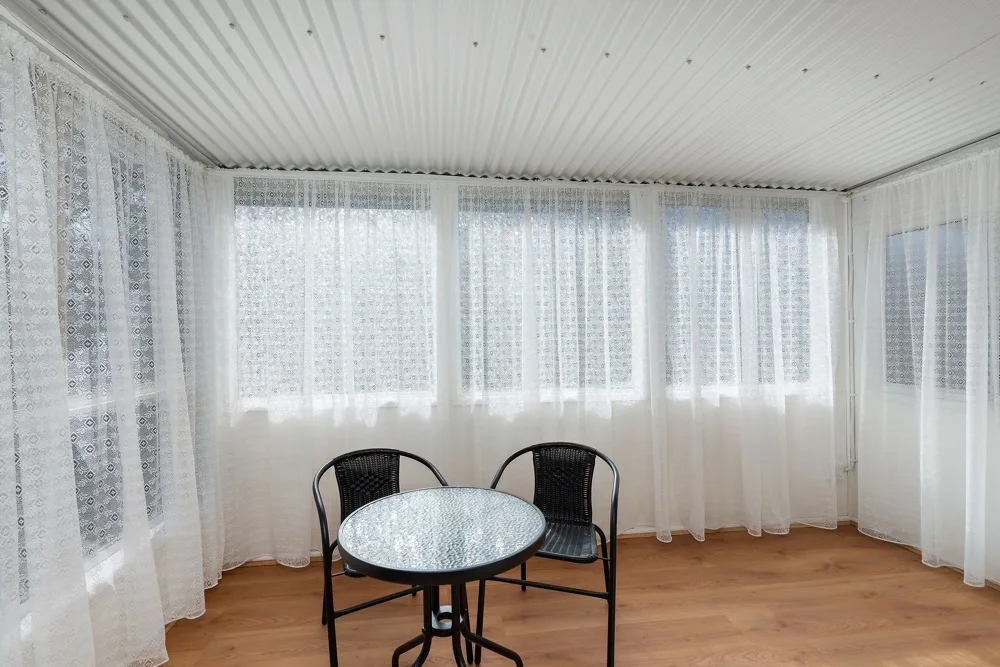 A bright enclosed sitting area with a small round table and lace curtains.