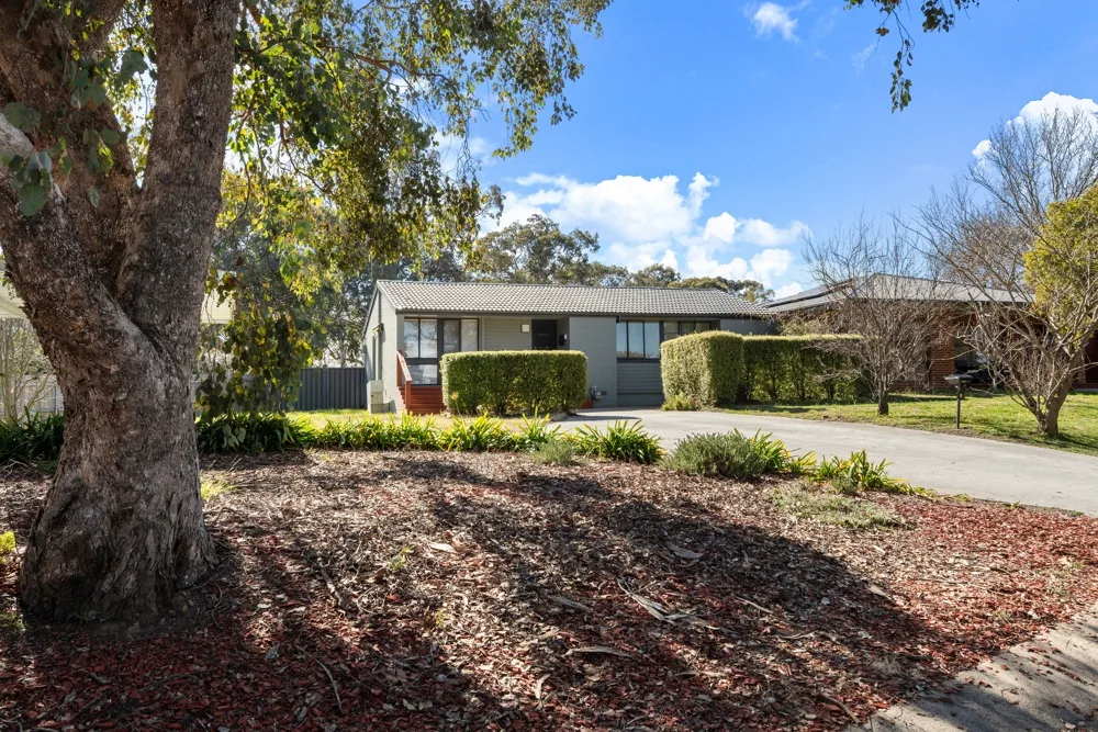 A single-story house stands beside a driveway, with trees and shrubs in front.
