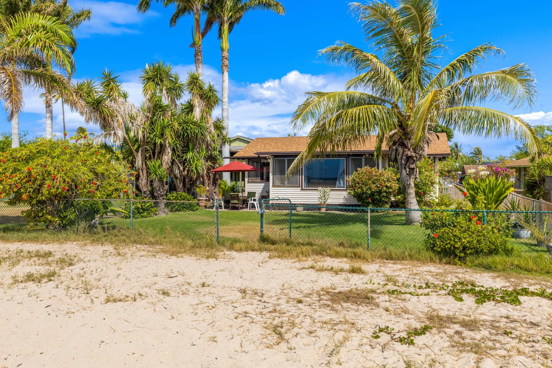 Una casa de una planta se encuentra detrás de una zona arenosa, rodeada de palmeras y arbustos.