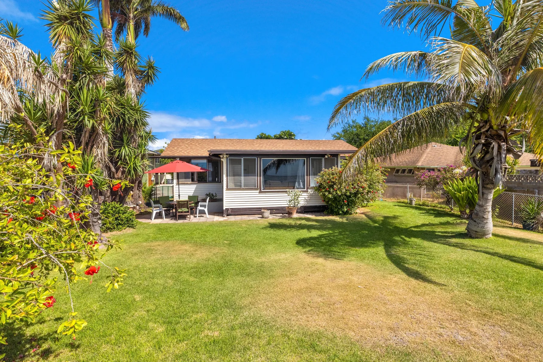 Una casa pequeña se encuentra al fondo de un césped soleado, con palmeras y asientos de patio.