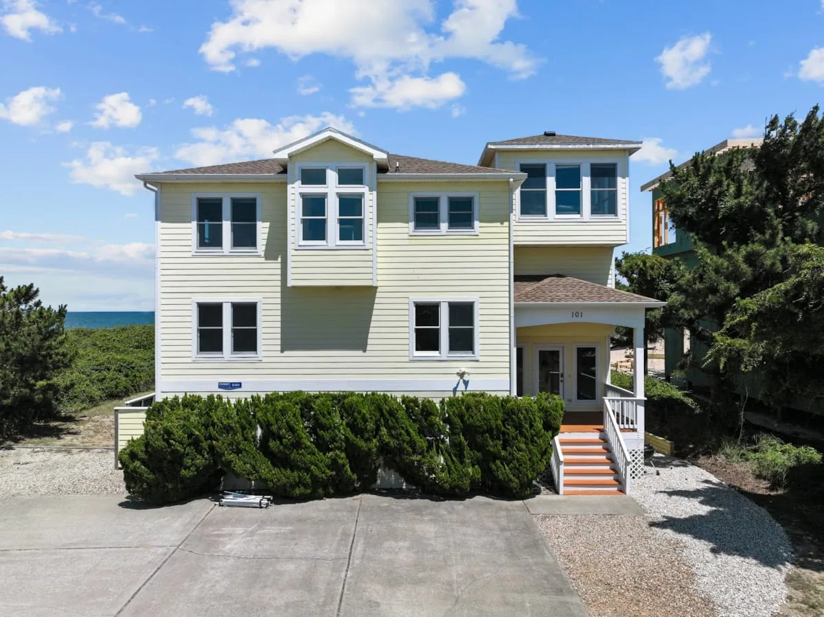 A pale two-story coastal house stands behind shrubs beside a paved driveway.