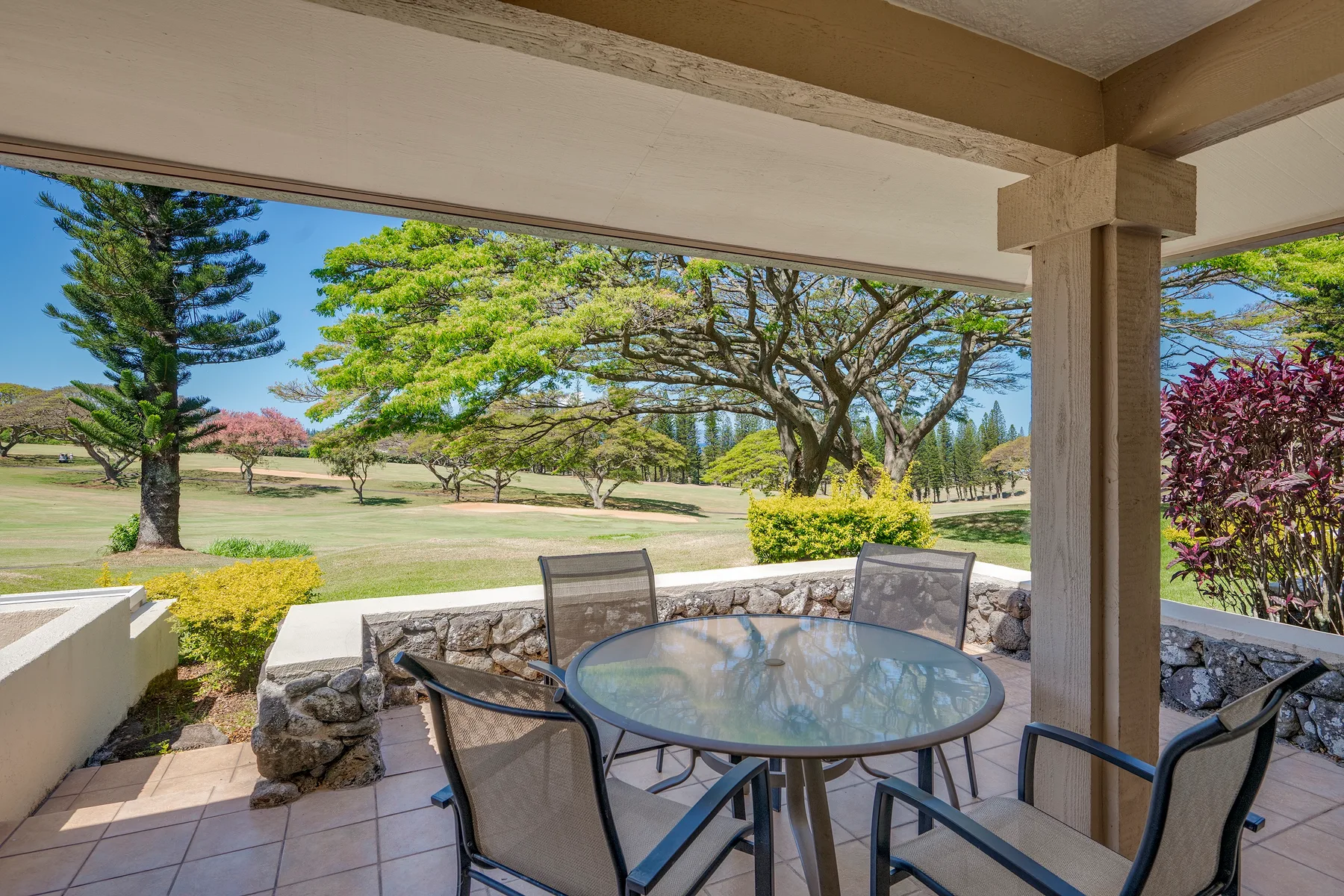 A covered patio has a round glass table and seating overlooking a landscaped garden.