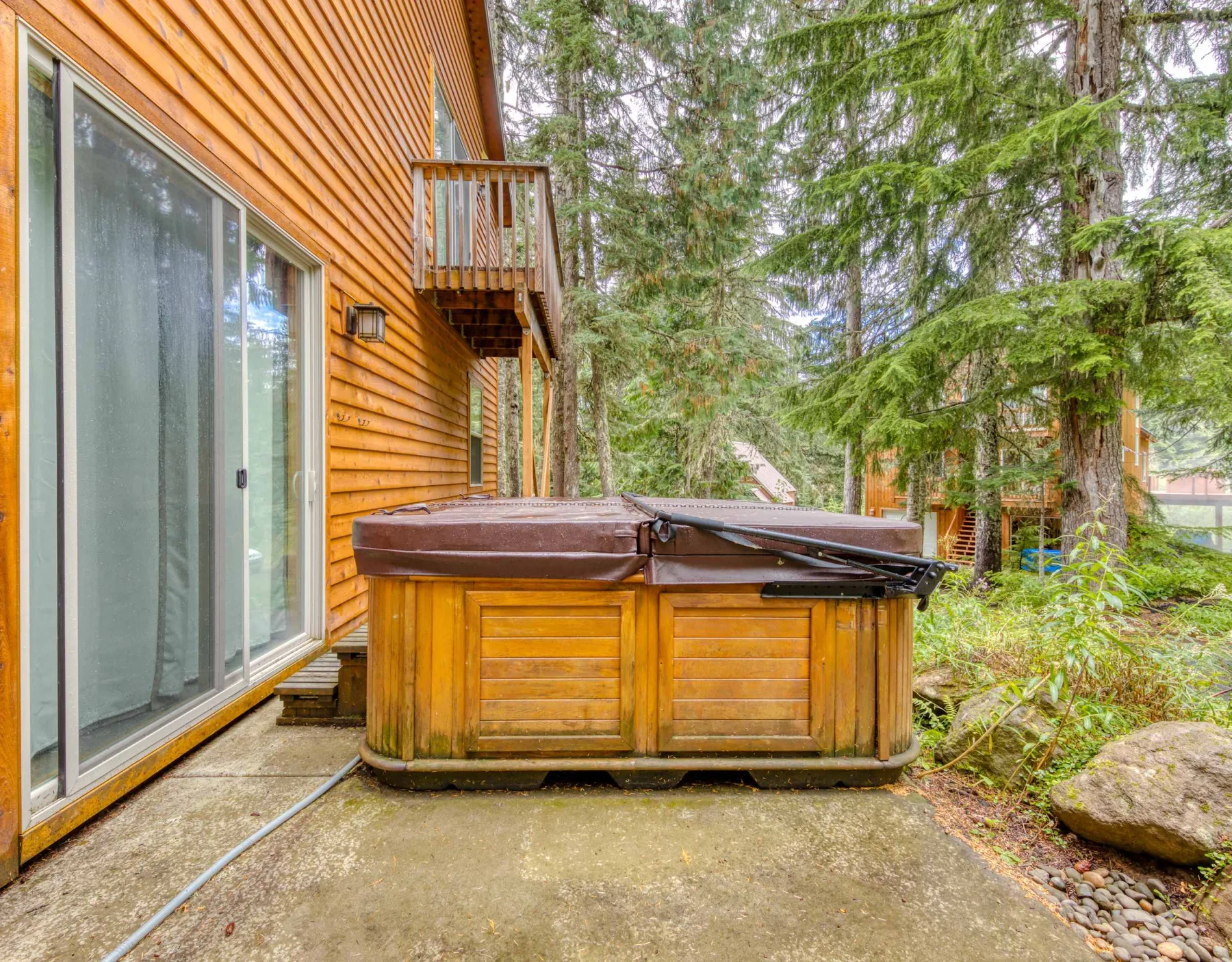 A wooden hot tub sits beside a house, surrounded by trees and greenery.