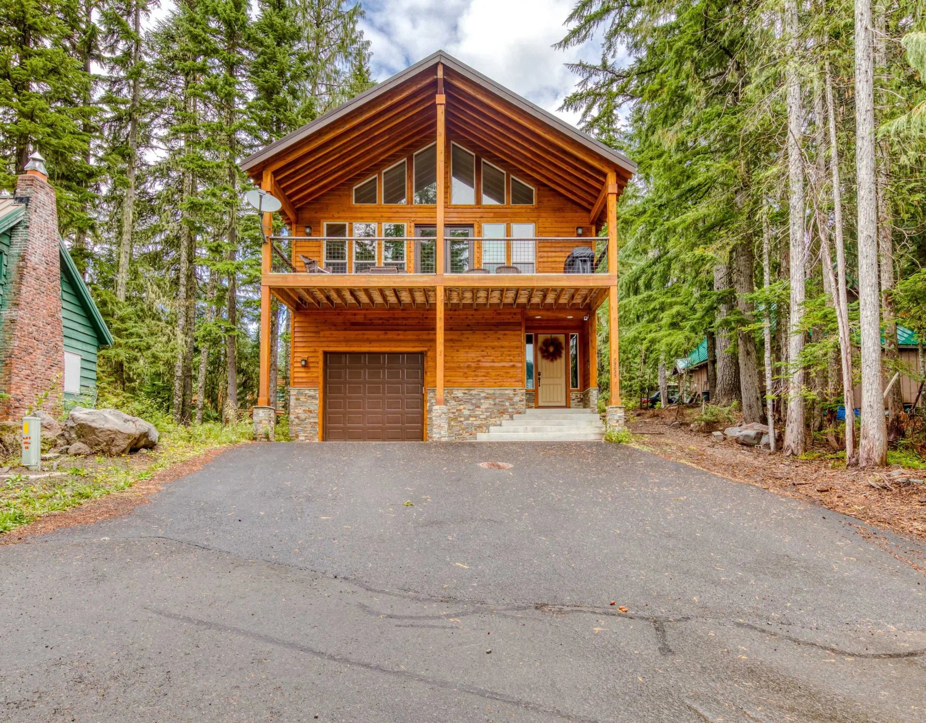 A wooden cabin stands among tall trees at the end of a paved driveway.