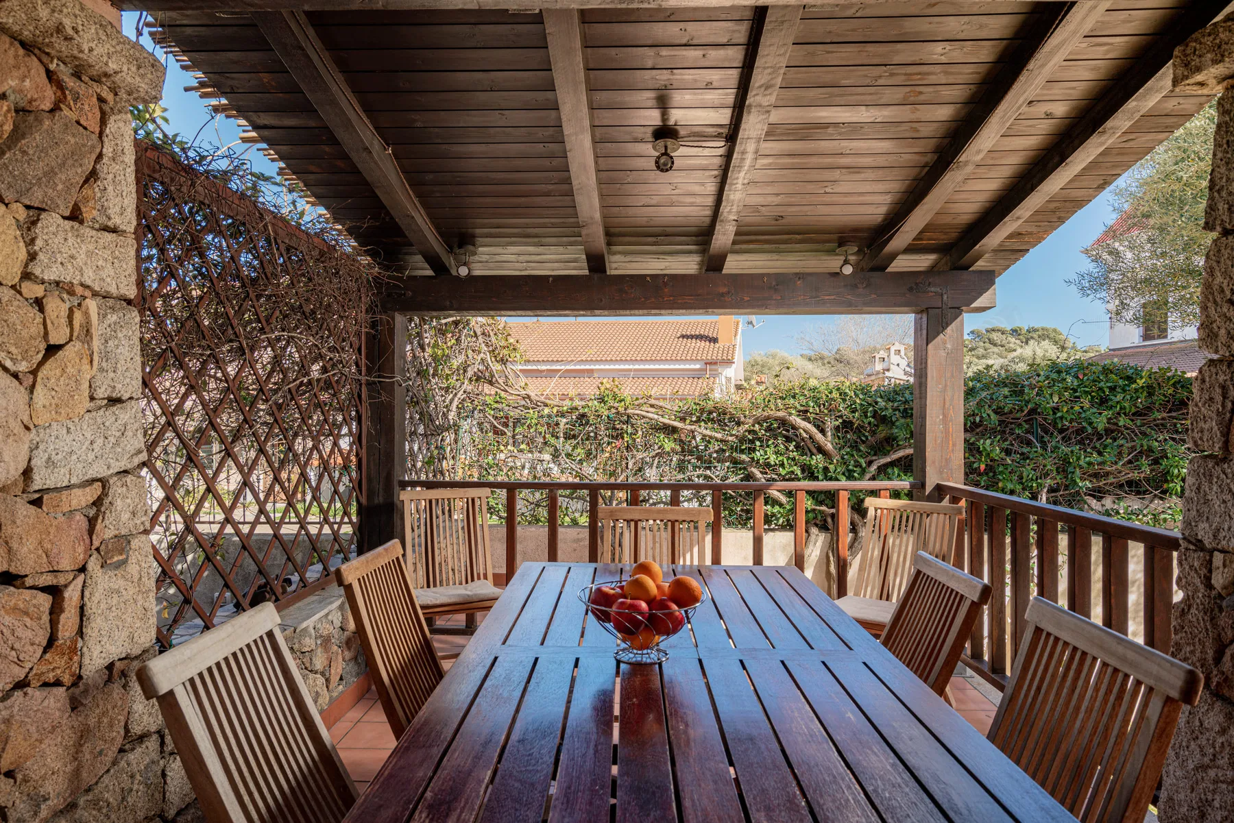 Comedor exterior cubierto con mesa y sillas de madera, junto a vegetación.