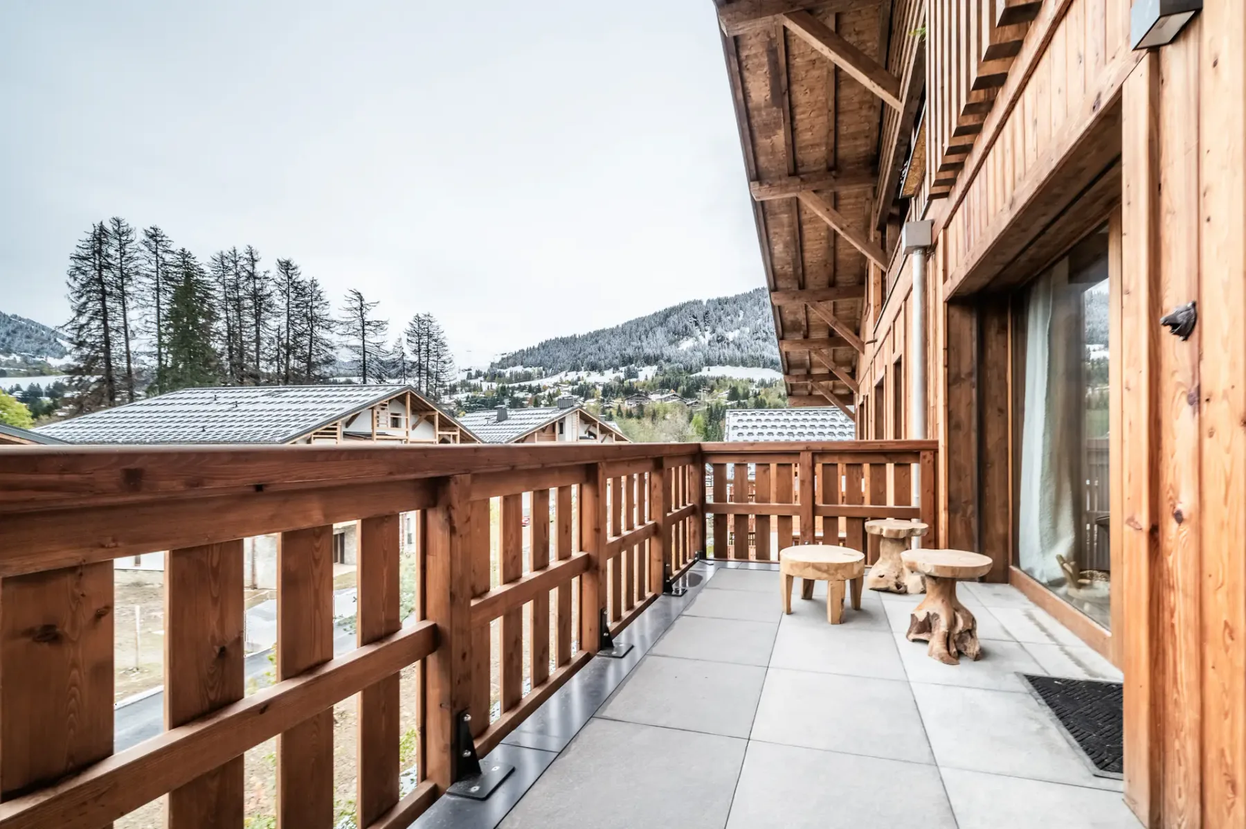 A wooden balcony with outdoor seating overlooks nearby rooftops and a wooded hillside.