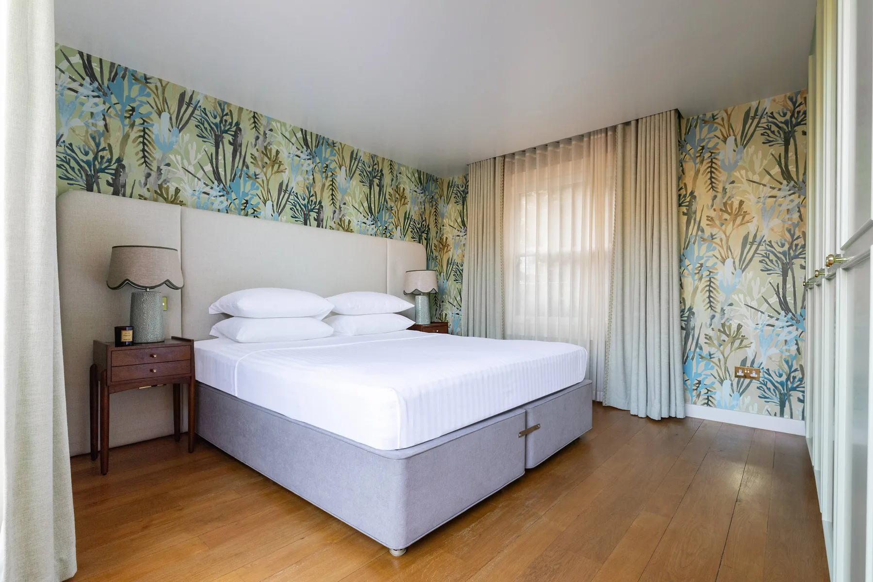 A bedroom with a neatly made bed and patterned botanical wallpaper.