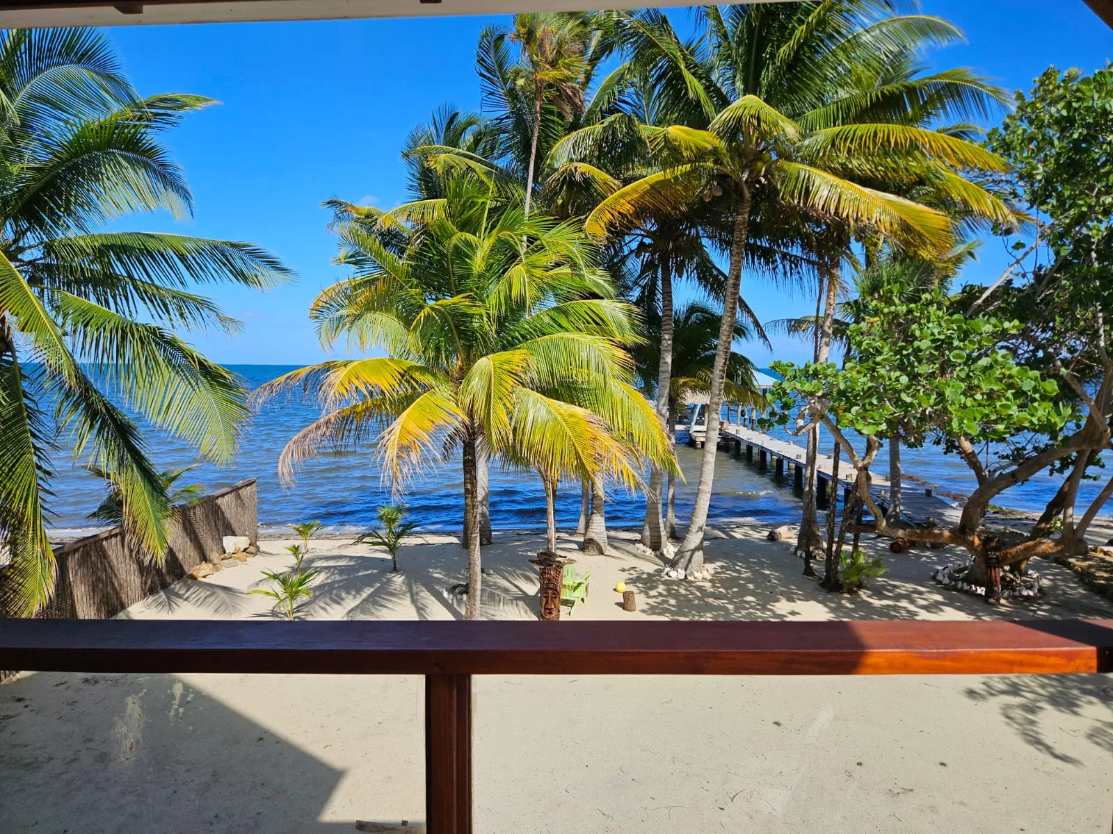 Una orilla tropical de arena con palmeras, agua azul y un muelle de madera.