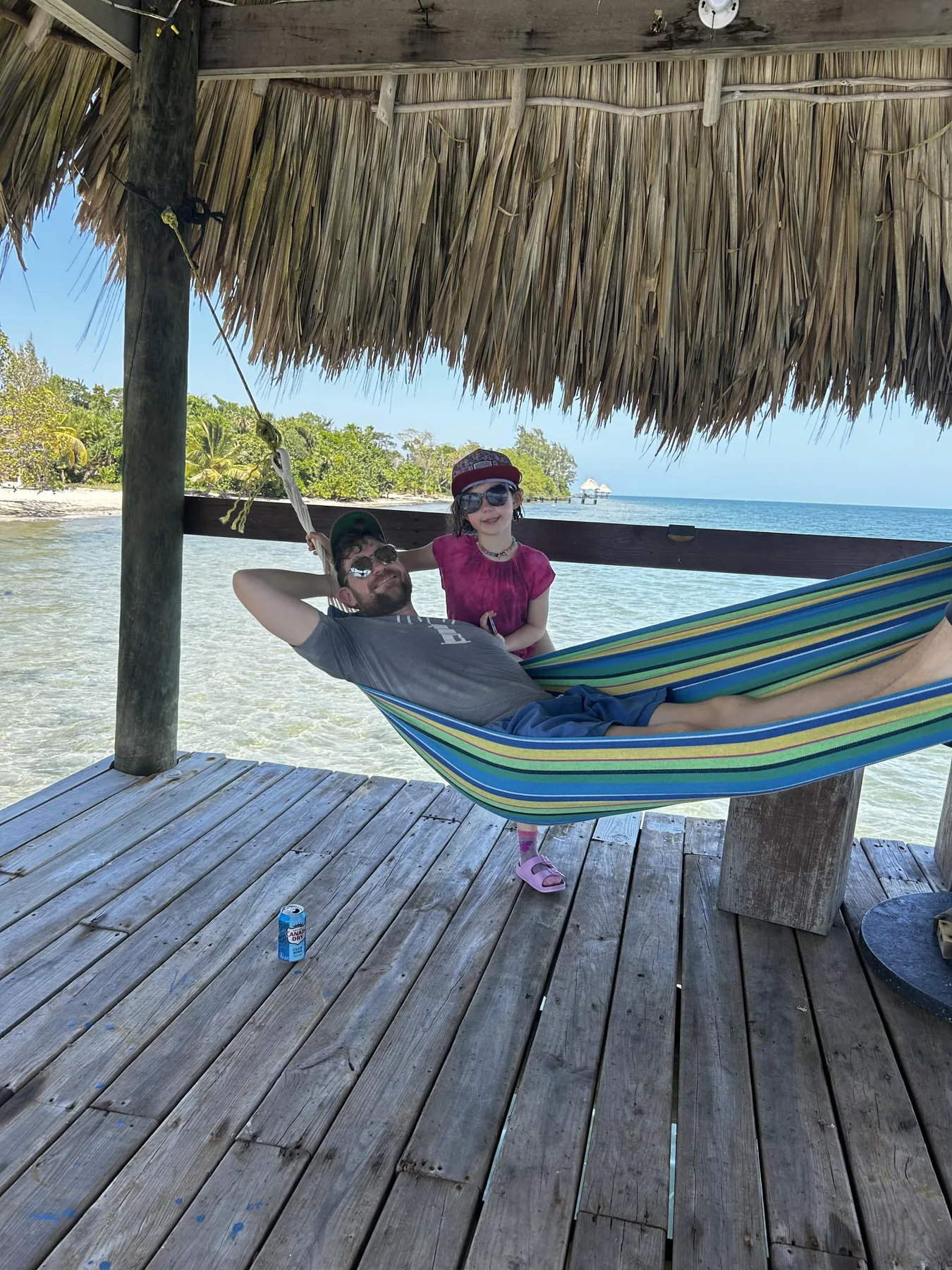 Un refugio de playa con techo de paja da al agua, con una hamaca y dos personas descansando.