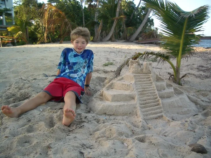 Un niño está sentado en una playa de arena junto a un castillo de arena detallado.