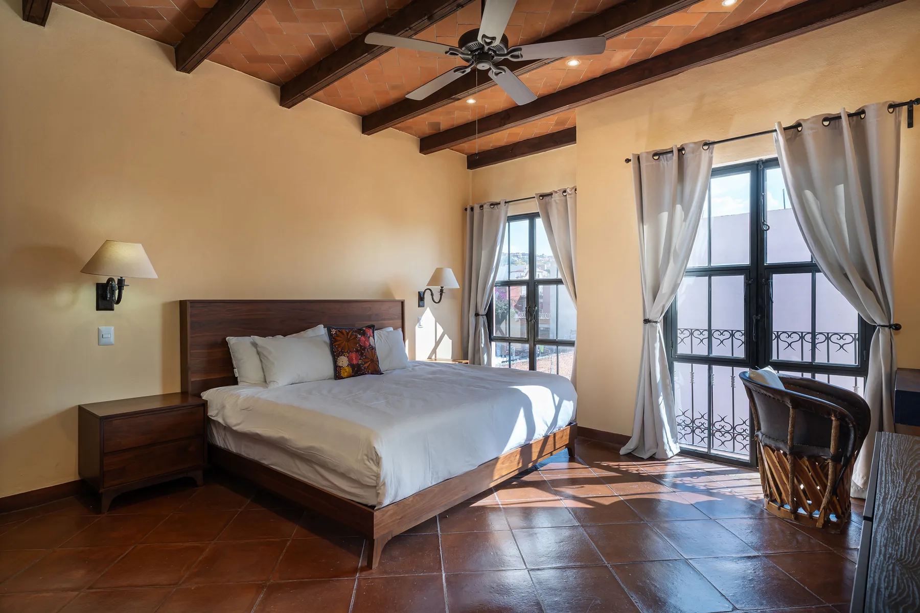 A bedroom with a large bed, exposed wooden ceiling beams, and tall curtained windows.
