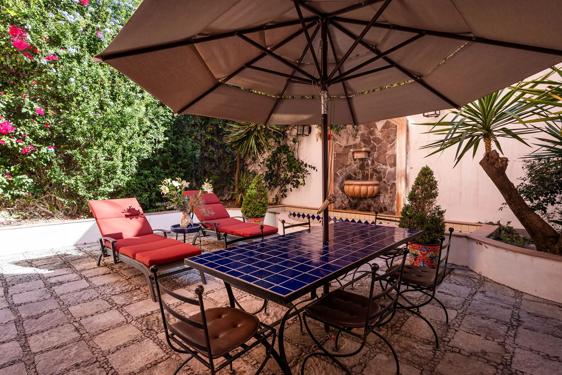 A garden patio has a tiled table beneath a large umbrella, surrounded by seating and plants.