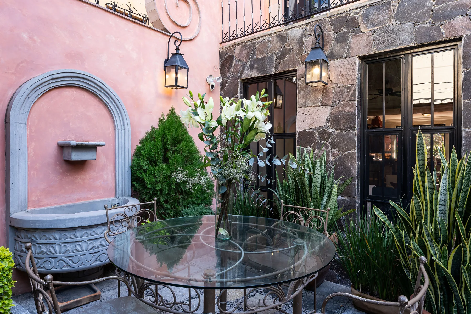 A small courtyard patio has a round glass table, seating, and potted plants.