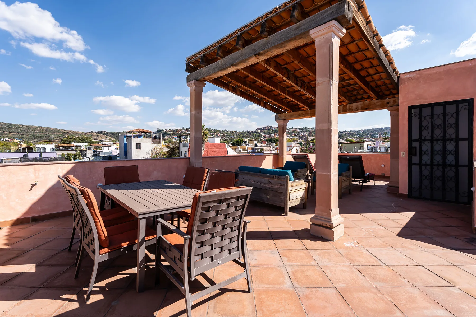 A rooftop terrace has outdoor seating beneath a wooden pergola, with a city view.