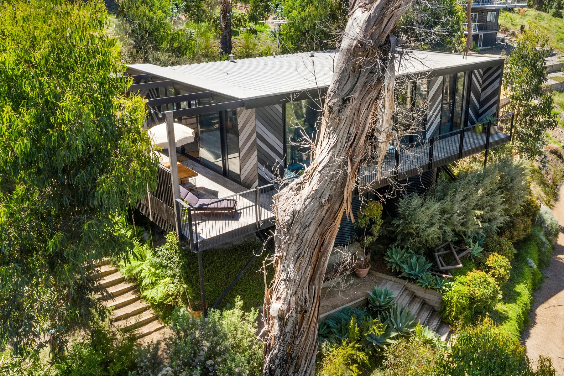 Una casa moderna con una amplia terraza está rodeada de árboles y vegetación ajardinada.
