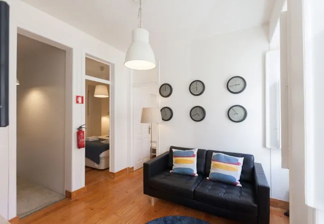 Una sala luminosa con un sofá oscuro y varios relojes en la pared.