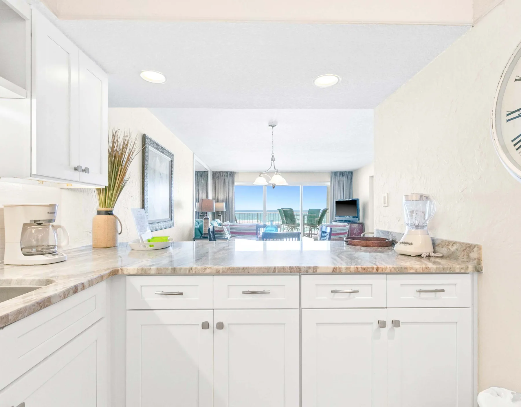 Una cocina luminosa con encimeras y gabinetes blancos se abre hacia una sala y vista al mar.