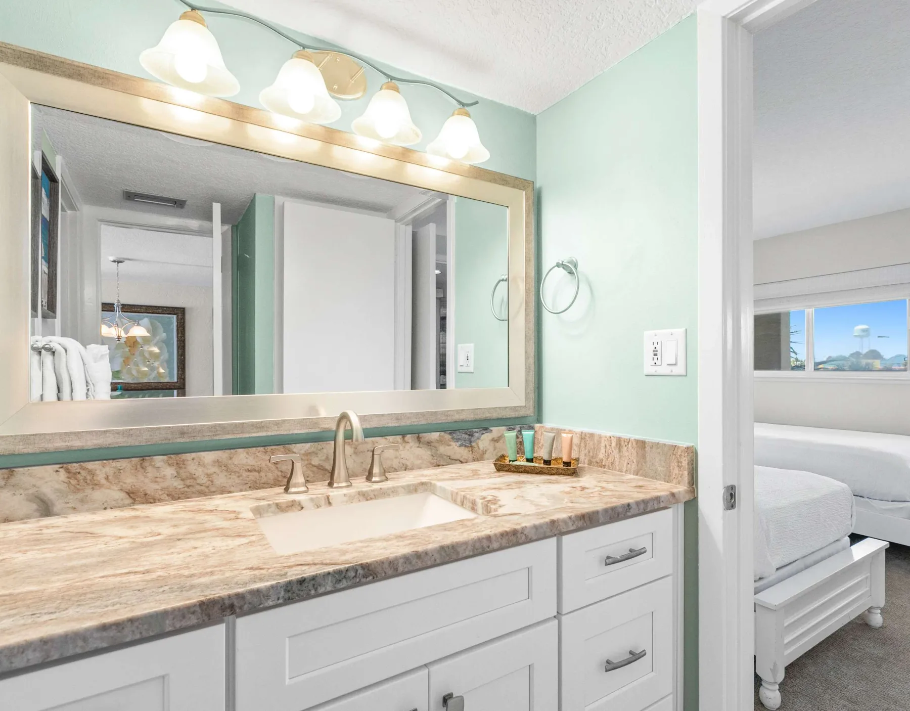 Un tocador de baño luminoso con un espejo amplio y un dormitorio visible por la puerta.