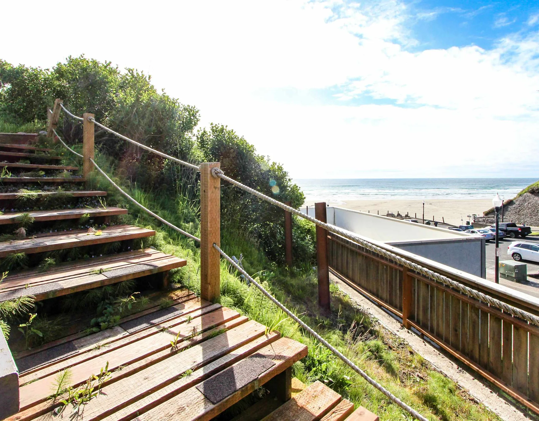 Wooden outdoor stairs descend beside a rope railing toward a beach and ocean.