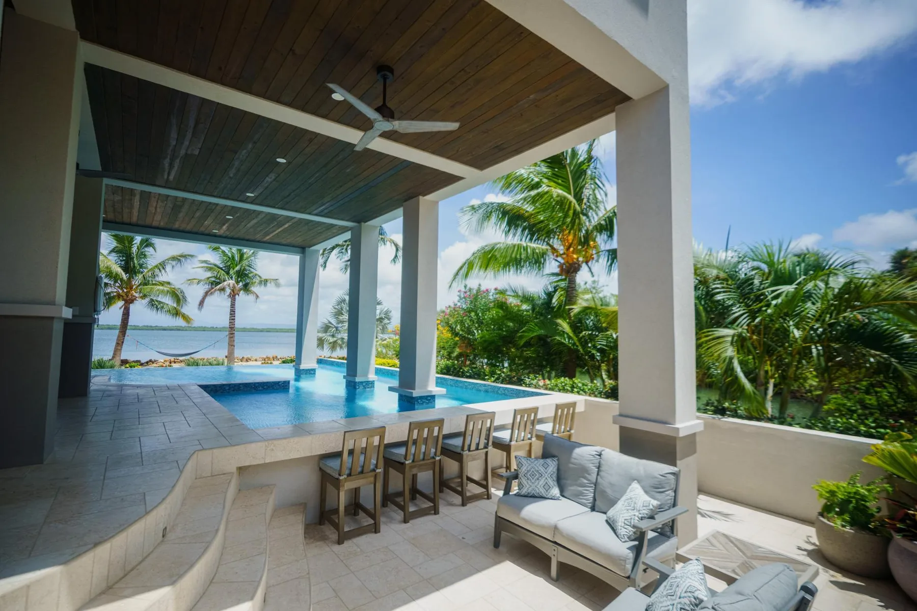 A covered patio with seating overlooks a pool and palm-lined ocean view.