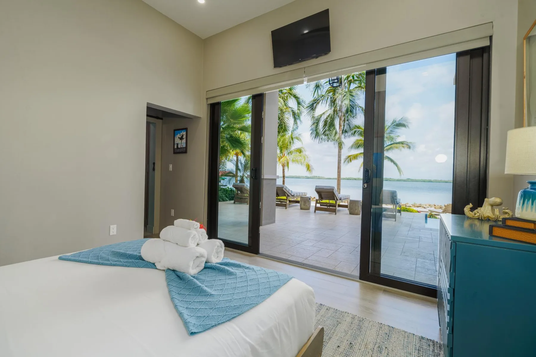 A bedroom opens through sliding glass doors to a patio with palm trees and water beyond.