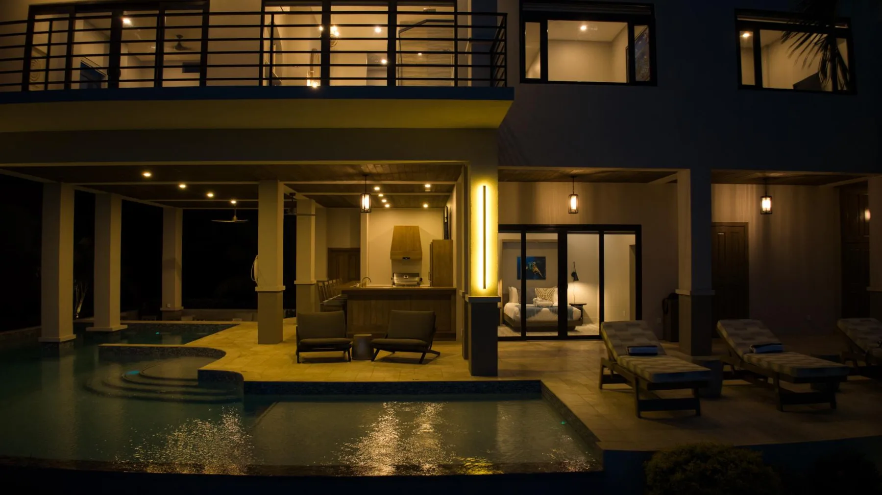 A modern house opens onto a lit patio with a pool at night.